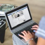 Mulher utiliza notebook apoiado no colo e acessa página do Facebook. Ela tem unhas pintadas de azul.