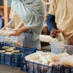 Sobre uma mesa, voluntários, uma mulher branca e um homem negro, organizam latas de alimentos e pães em caixas de plástico para transporte